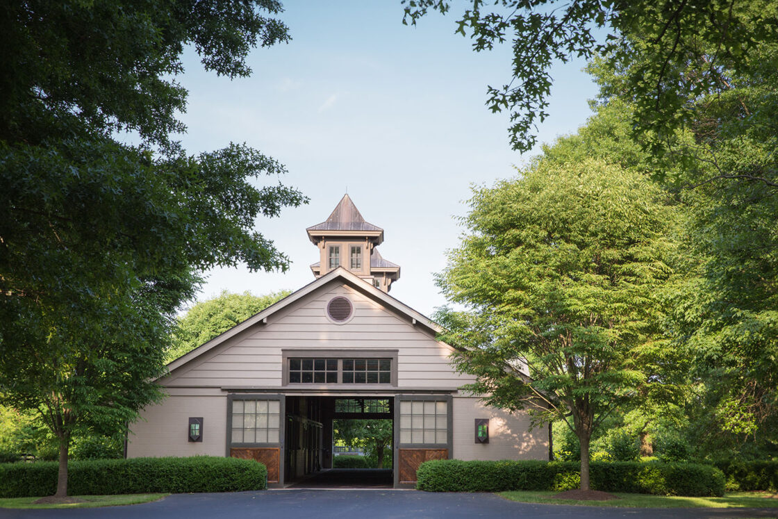 custom equine facility sitting beneath a clear sky with multiple trees and plans surrounding it while its front doors are open