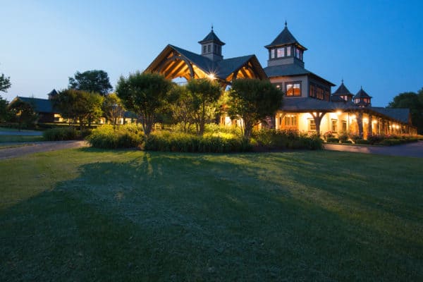 A large, elegant luxury horse barn with peaked roofs and glowing lights is surrounded by trees and a neatly manicured lawn at dusk. The clear blue sky enhances the peaceful, inviting atmosphere.