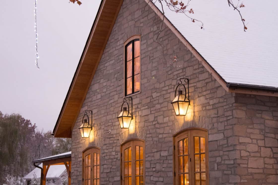 A stone luxury horse barn with tall wooden doors and warm glowing lanterns, surrounded by snow and bare tree branches, on a winter evening.