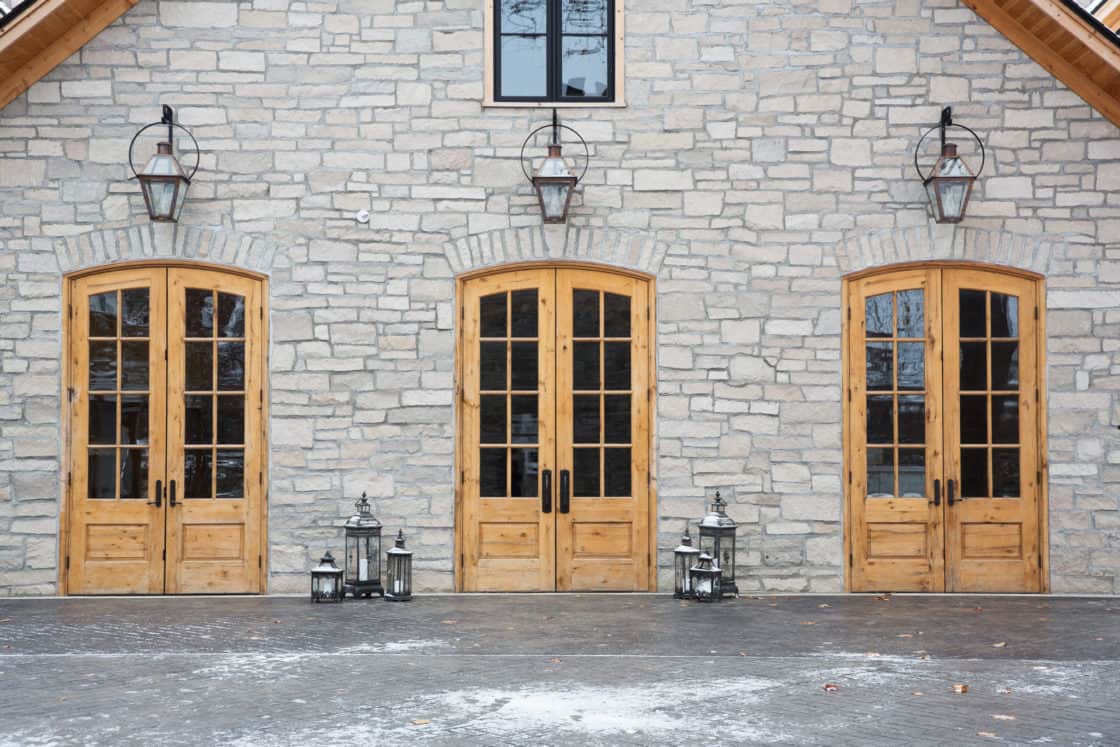Three wooden double doors set in a light stone bank barn wall, each topped with a glass transom and outdoor lanterns. Several metal lanterns are grouped on the snow-dusted wooden deck, adding rustic charm.