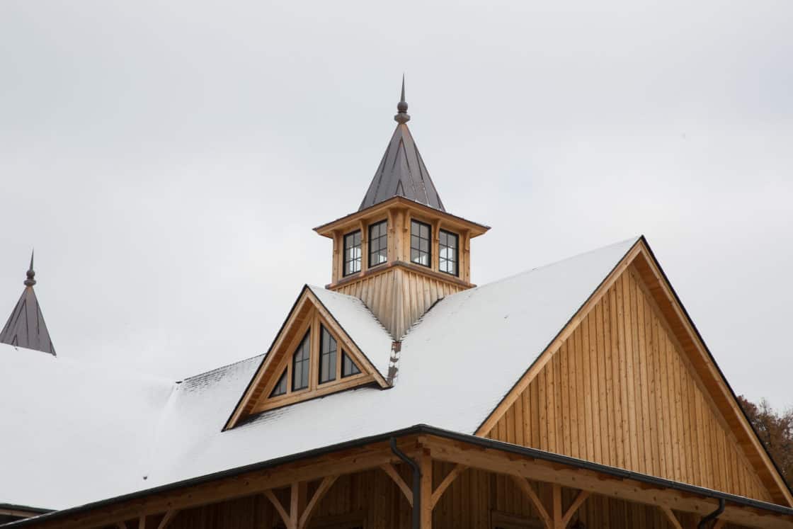 A wooden bank barn with snow on its roof features a small tower with windows and a pointed metal steeple. The sky above is overcast.