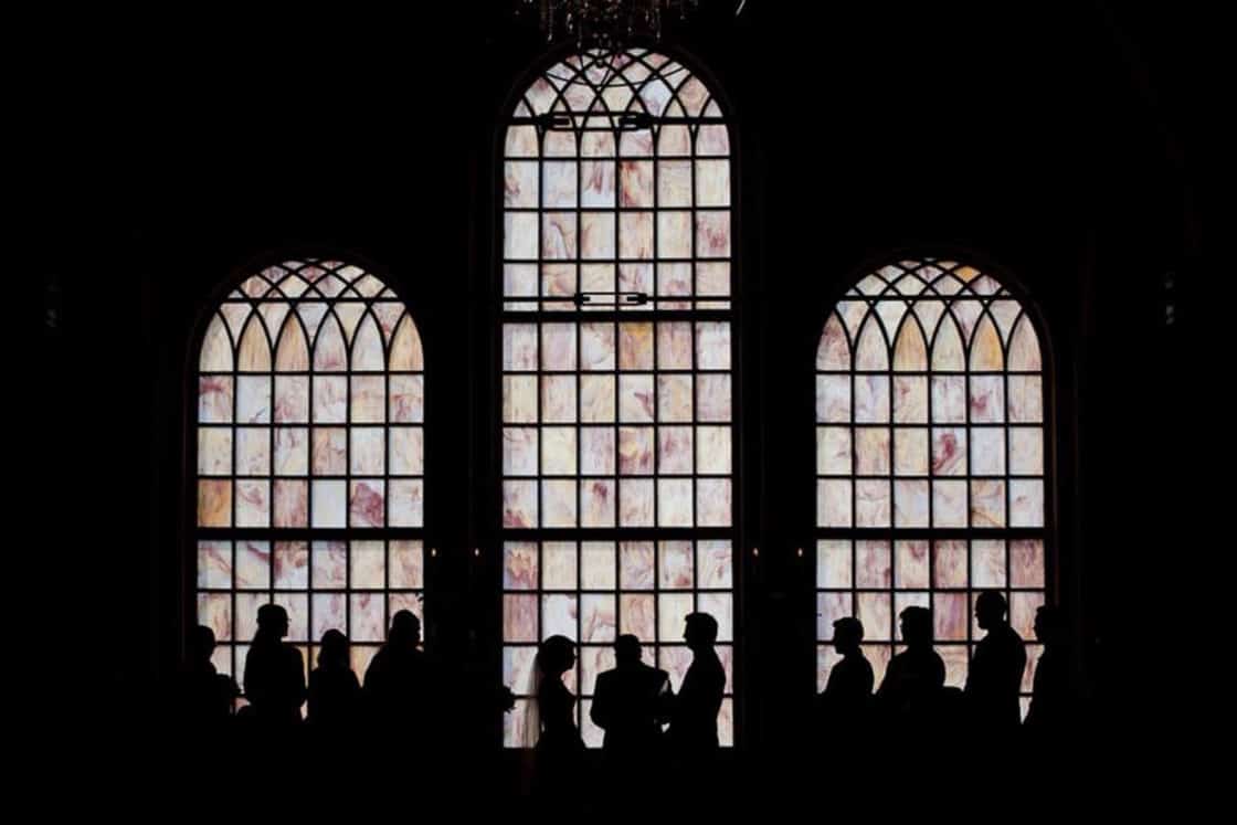 Silhouettes of people, possibly at a wedding, stand in front of large arched stained-glass windows inside a dark luxury home, with light softly filtering through the glass.