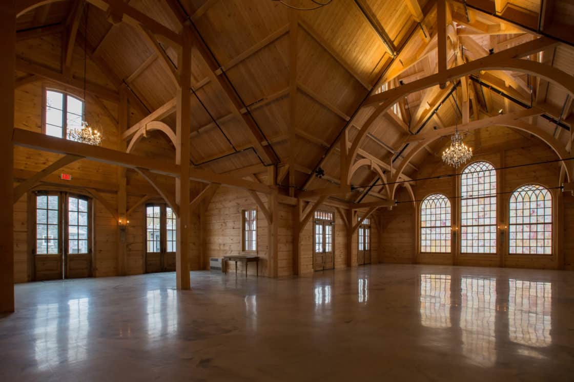 A spacious, empty wooden hall with high vaulted ceilings, large arched windows, exposed beams, and chandeliers, this bank barn lets in natural light and creates a warm, rustic atmosphere reminiscent of a luxury home.