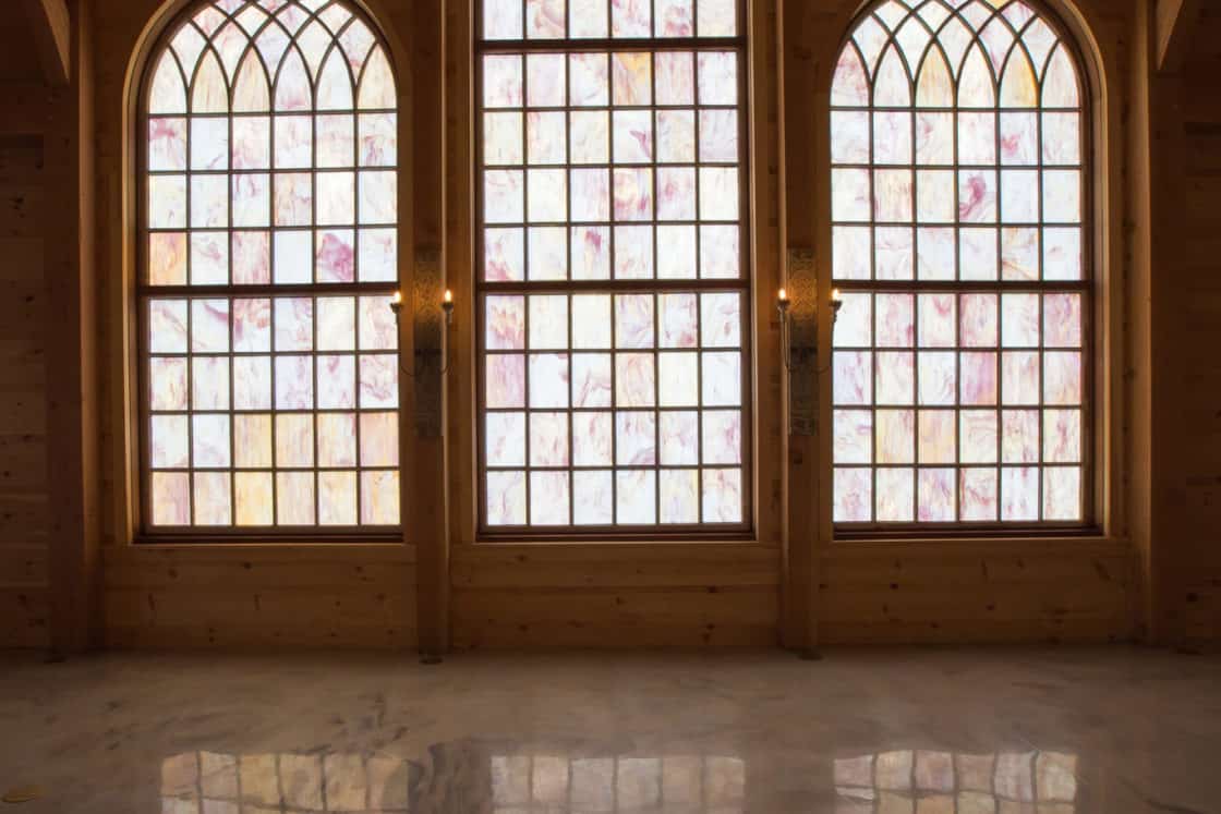 Three tall arched windows with stained glass panels grace the wooden wall of this luxury home, casting a faint colorful light onto the polished marble floor of an empty, warmly lit room.