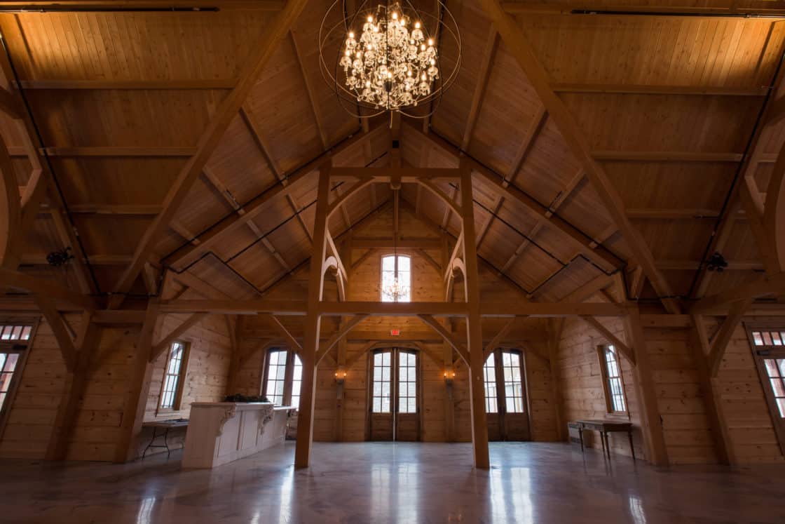 Spacious wooden party barn with high, vaulted ceilings, exposed beams, large windows, and a central chandelier; natural light fills the room, and there are tables along the walls.
