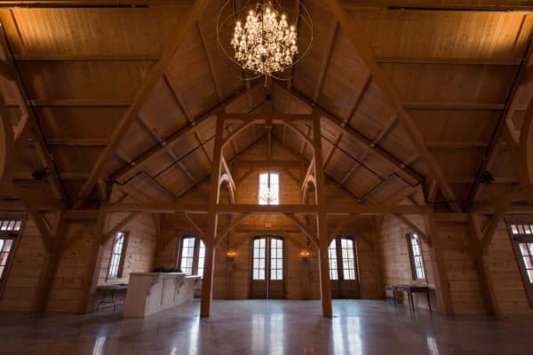 Spacious wooden party barn with high, vaulted ceilings, exposed beams, large windows, and a central chandelier; natural light fills the room, and there are tables along the walls.