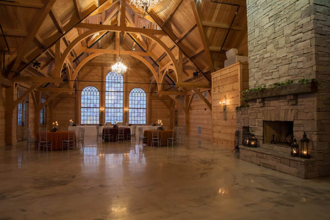 A spacious party barn event hall with high wooden beams, large arched windows, chandeliers, round tables with chairs, and a stone fireplace decorated with candles and lanterns.
