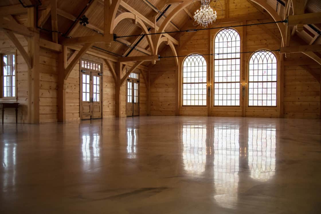A spacious, empty wooden hall with arched windows, high vaulted ceiling, exposed beams, and a chandelier—perfect as a luxury horse barn or party barn. Sunlight streams through tall windows, reflecting on the polished floor.