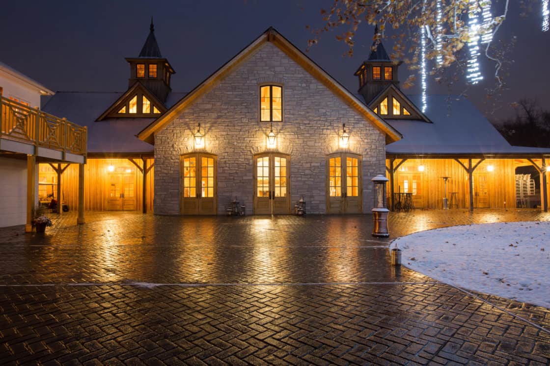 A warmly lit stone and wood party barn with tall peaked roofs and glowing windows stands on a wet, brick courtyard at night, with a dusting of snow on the ground and holiday lights overhead.