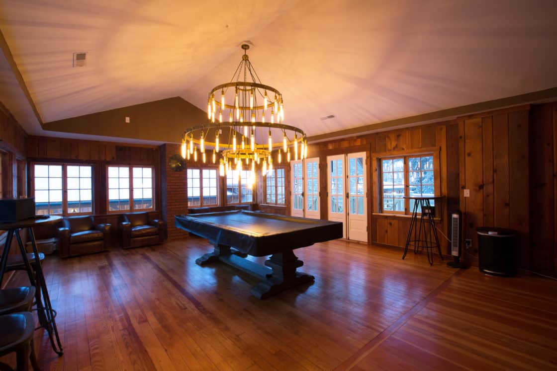 A spacious wooden game room in this luxury home features a large pool table under a grand chandelier, surrounded by windows, French doors, armchairs, and warm wood-paneled walls and floors.