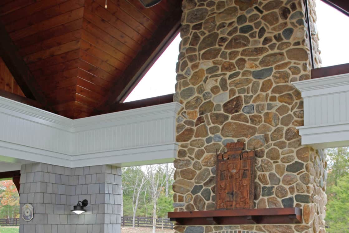 A tall stone fireplace with a wooden mantle stands under a vaulted wooden ceiling in an outdoor covered patio of a luxury home, featuring a round table and chairs. Green trees are visible outside.