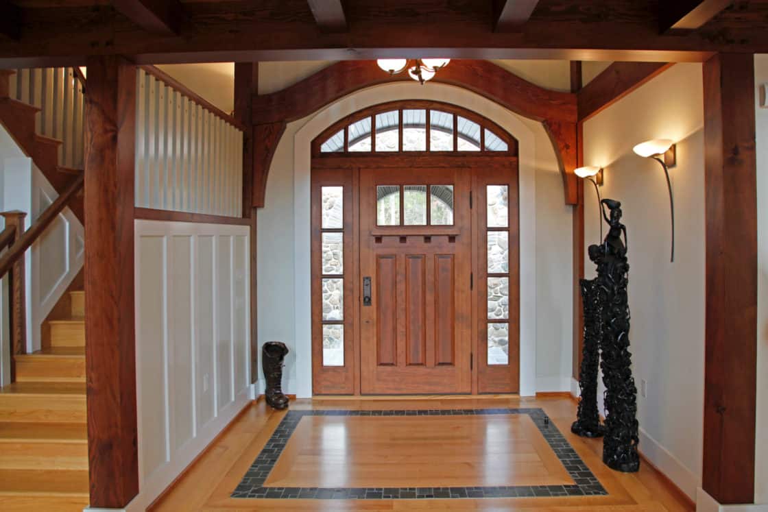 A wooden front door with glass panels is framed by sidelights and an arched window, echoing the grandeur of a luxury home. The entryway features wood floors, a staircase on the left, white walls with wainscoting, and abstract black sculptures.