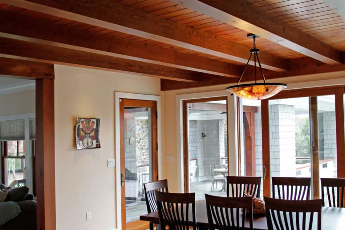 A luxury home dining room with a large wooden table and eight dark chairs, light wood flooring, exposed ceiling beams, a modern chandelier, and large windows filling the space with natural light.