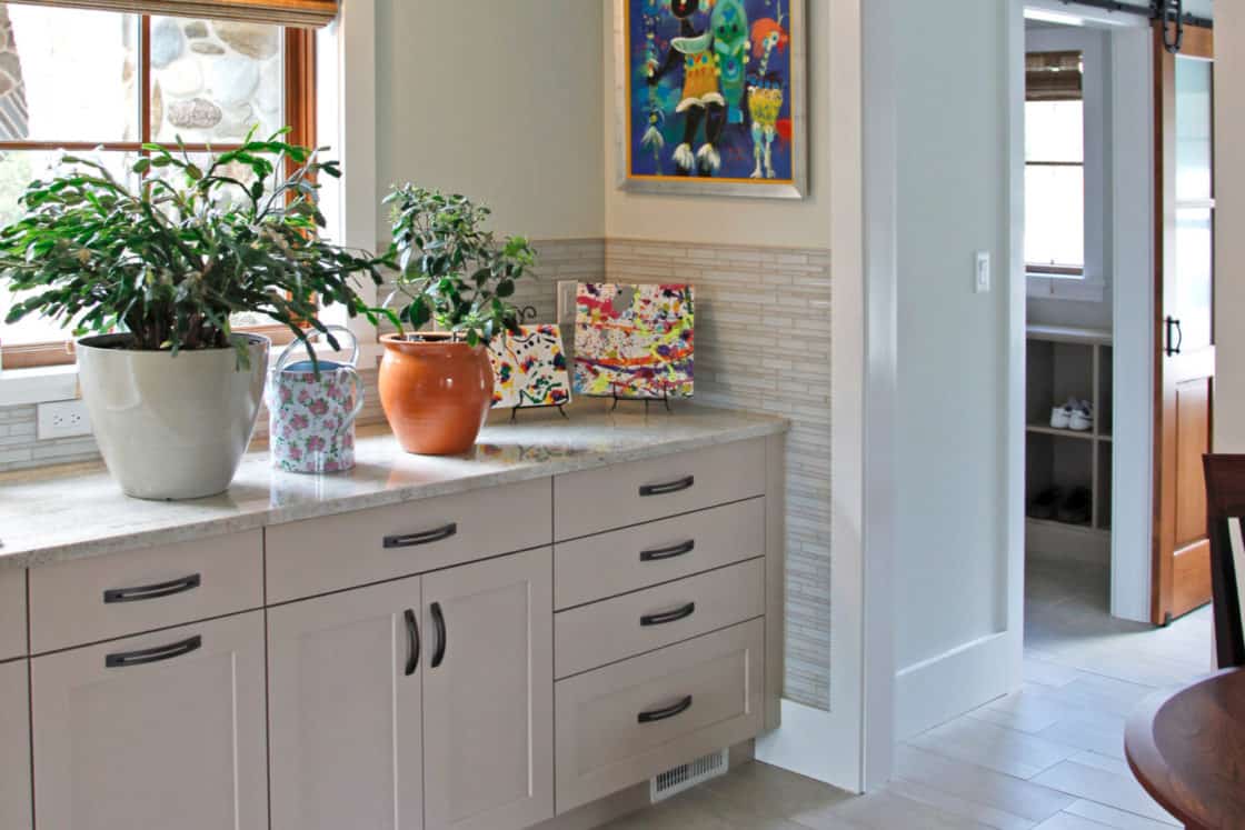 A bright kitchen corner with light cabinets, potted plants, and colorful artwork on the wall. A window with a bamboo shade lets in natural light, adding charm to this luxury home, where a sliding barn door hints at modern elegance in the hallway.
