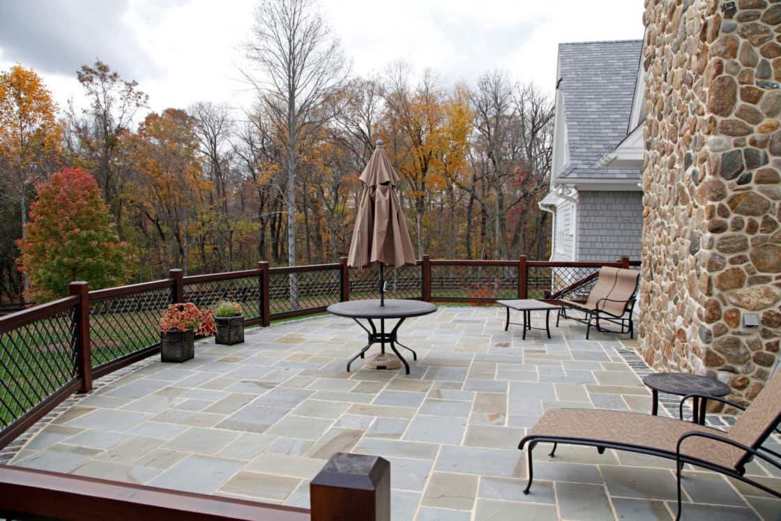 A spacious stone patio at this luxury home features lounge chairs, a round table, a closed umbrella, potted plants, and a wooden railing. Trees with autumn foliage create a stunning backdrop for relaxing or entertaining.