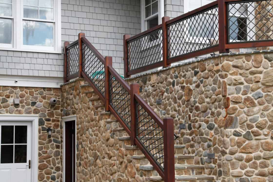 A stone house with an exterior staircase featuring a dark wood and metal railing leads to a patio. Reminiscent of a classic bank barn, it boasts grey shingles, white trim, large windows, and landscaped plants in the yard.
