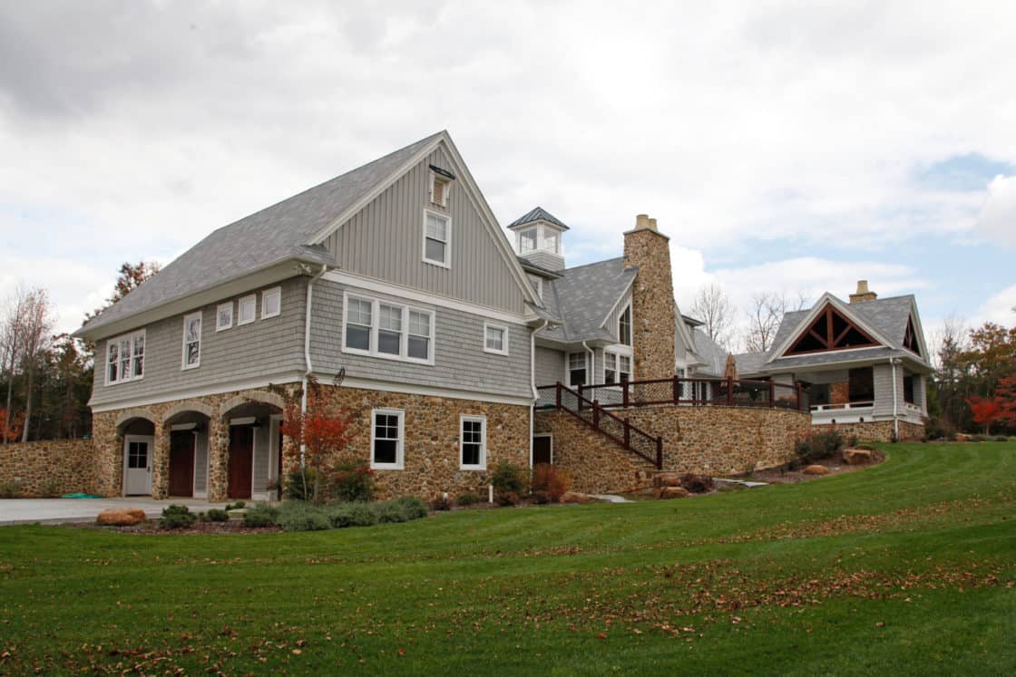 A large two-story luxury home with gray siding, stone accents, arched garage doors, and a spacious lawn. A stone stairway leads to a porch, and a covered outdoor area is attached to the right side. Trees are visible in the background.