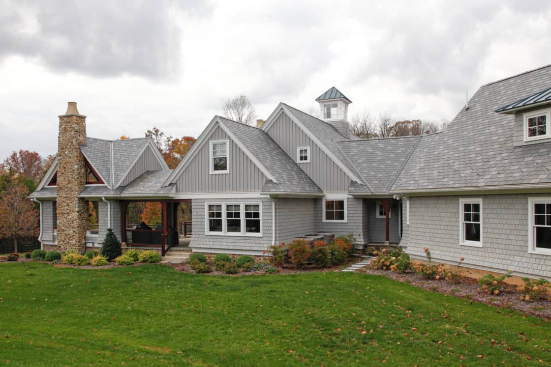 A large gray luxury home with gabled roofs, stone chimney, white trim, and a cupola, surrounded by green lawn and landscaped bushes under a cloudy sky.