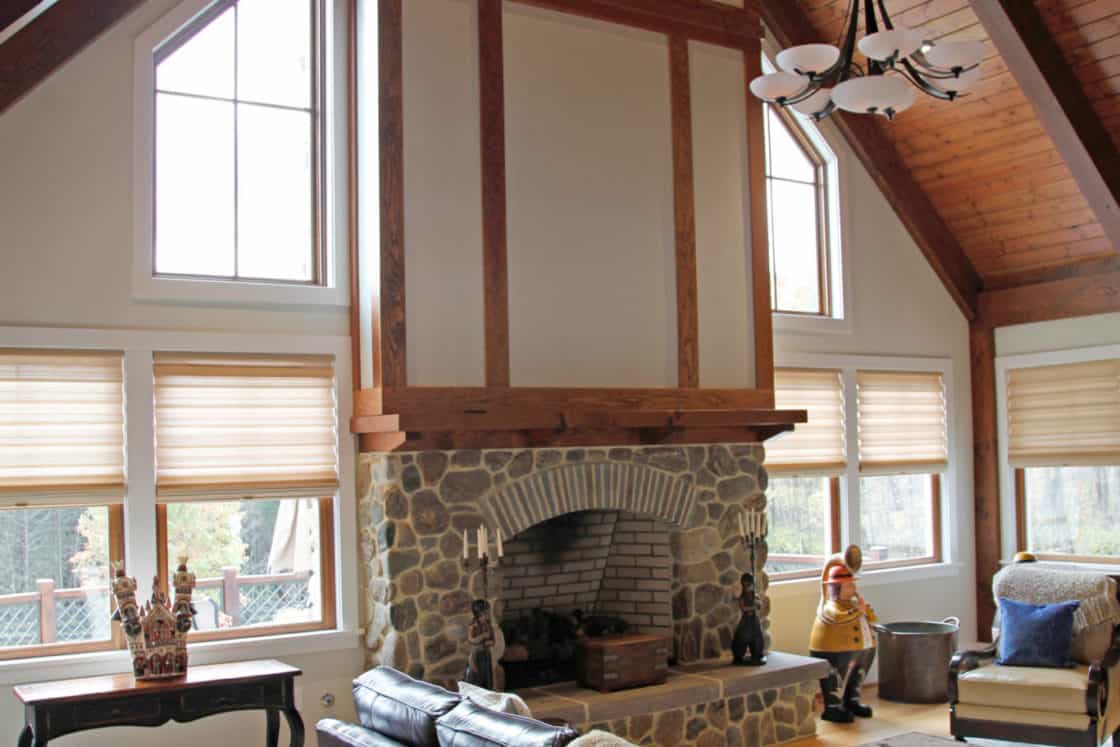 A spacious living room in this luxury home features a tall stone fireplace, wood beams, large windows, leather sofas, a chandelier, and wooden floors. Bright natural light fills the stylish space, reminiscent of a refined party barn.