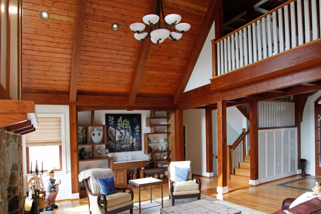 A spacious living room with exposed wooden beams, high vaulted ceiling, a chandelier, two armchairs and a sofa, wooden floors, a rug, and a staircase with white railings evoke the rustic charm of a luxury horse barn.