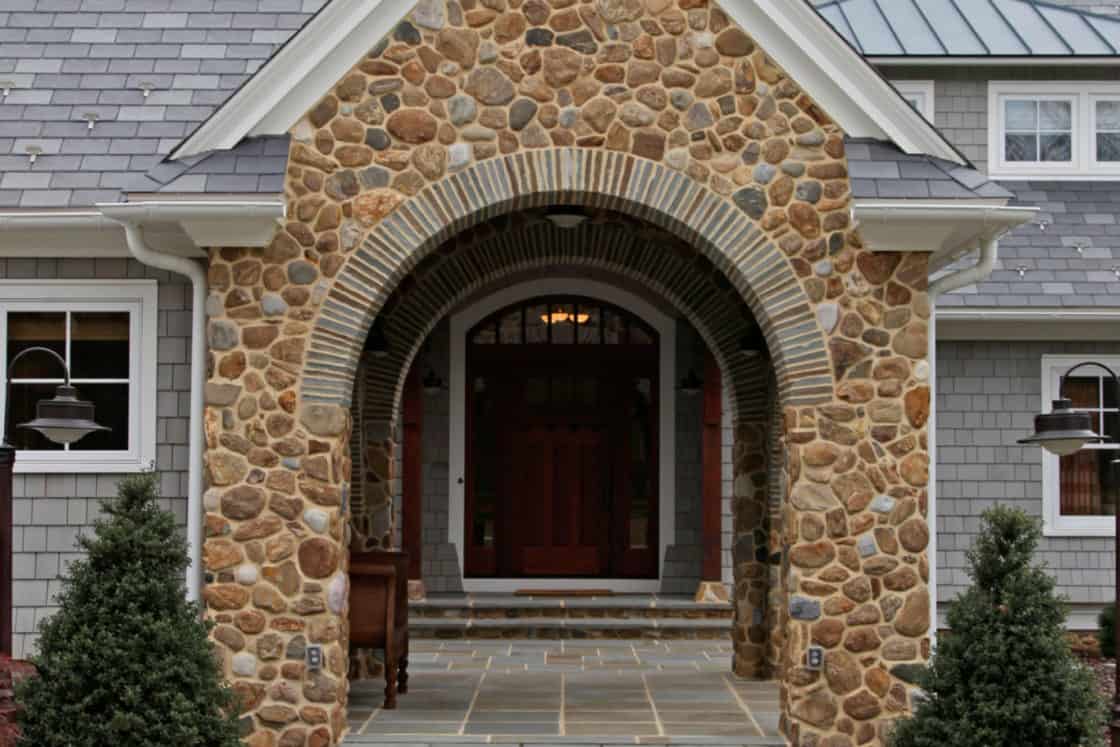 A stone archway with a covered entrance leads to the wooden front door of this luxury home, flanked by small evergreen shrubs and a stone walkway. The house features gray shingles and a metal roof.