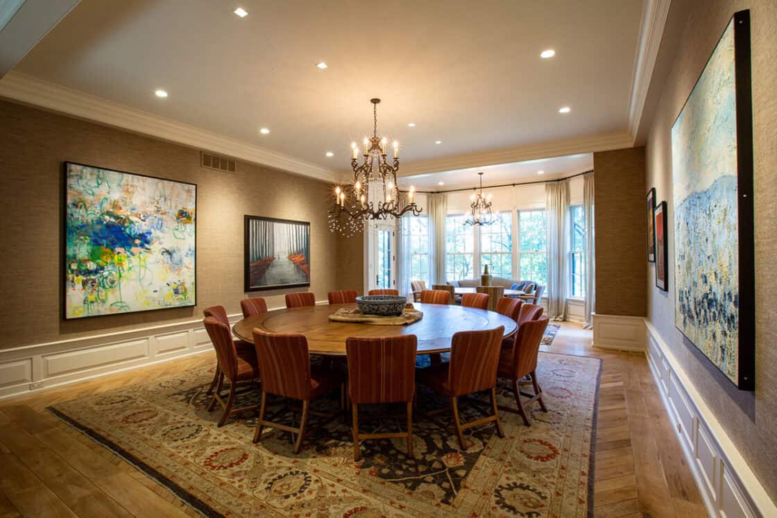 A spacious dining room reminiscent of a luxury horse barn, featuring a large round wooden table surrounded by red cushioned chairs, modern abstract artwork, two chandeliers, and large windows letting in natural light.