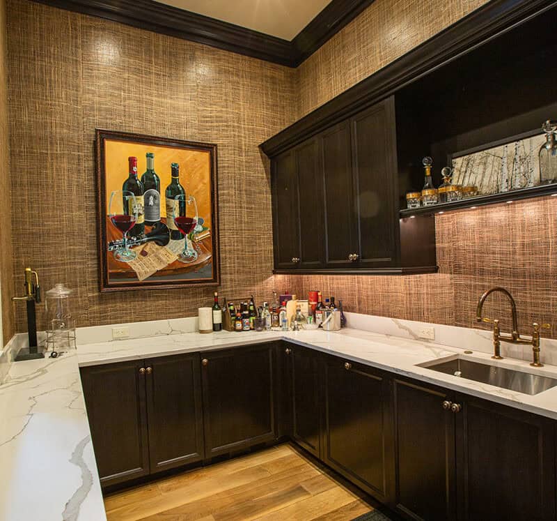 A luxury home bar with dark cabinets, marble countertops, and a sink. Bottles and glasses are arranged on the counter. Art featuring wine bottles and glasses hangs on a textured brown wall. Warm lighting brightens the space.