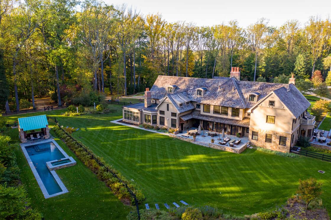 Aerial view of a luxury home with a spacious backyard, patio seating, and a rectangular swimming pool, surrounded by lush green lawn, tall trees, and a nearby luxury horse barn.