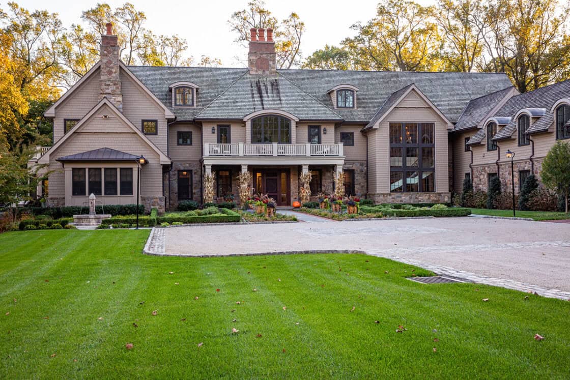 Large two-story luxury home with stone and wood exterior, multiple chimneys, arched windows, a central balcony, and a spacious driveway surrounded by green lawn and landscaped plants. Trees are visible in the background.