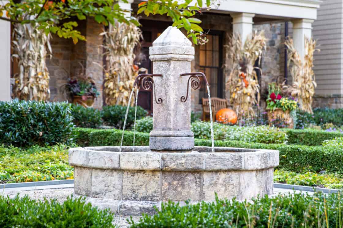 A stone fountain with water streams stands in a landscaped garden with green shrubs, in front of a luxury home decorated with cornstalks and pumpkins near the entrance.