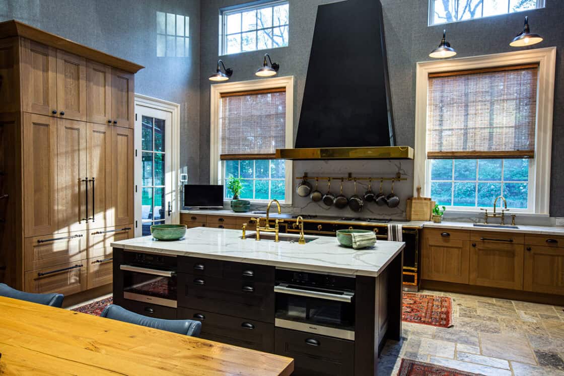 Spacious kitchen in a luxury home with wooden cabinets, a large black range hood, white marble island with brass faucets, hanging pots, windows with bamboo shades, and natural light illuminating the space.