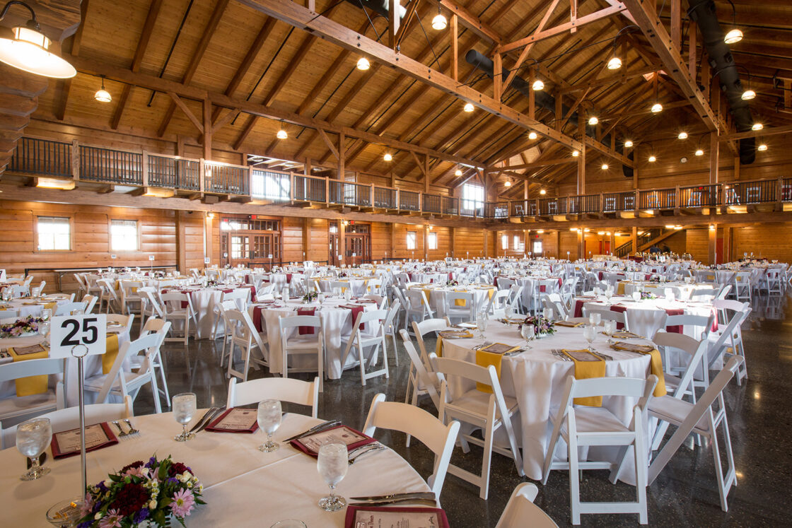 A large rustic banquet hall reminiscent of a party barn with wooden beams and high ceilings, set up for an event with round tables draped in white cloths, elegant centerpieces, glasses, and menus. White chairs surround the tables; table 25 is visible.