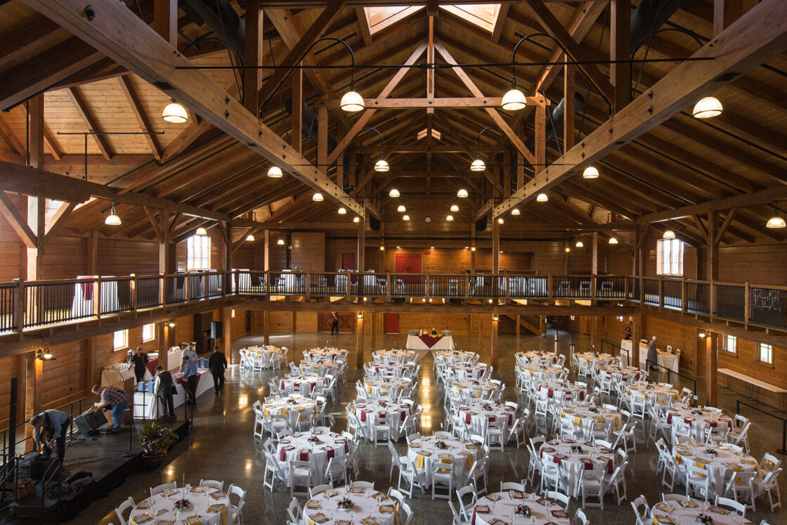 Large rustic banquet hall styled like a luxury horse barn, with exposed wooden beams, round tables set with white tablecloths and red napkins, a stage on the left, and an upper balcony overlooking the main floor.