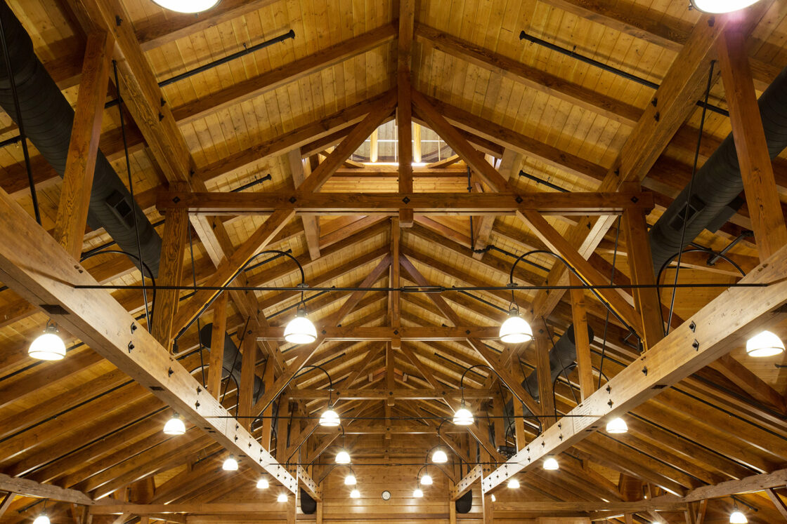 Wooden ceiling with exposed beams and trusses, reminiscent of a party barn, features multiple hanging round lights and black ventilation ducts. Warm lighting highlights the natural wood texture and the geometric structure of the roof.