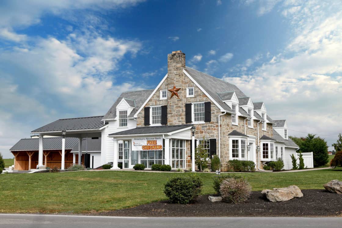 A large, two-story luxury home with stone and white siding, a gray gabled roof, and a front porch. A sign reading “Free Cheesecakes” hangs above the front windows. Green lawn and shrubs sit in front under a partly cloudy sky.