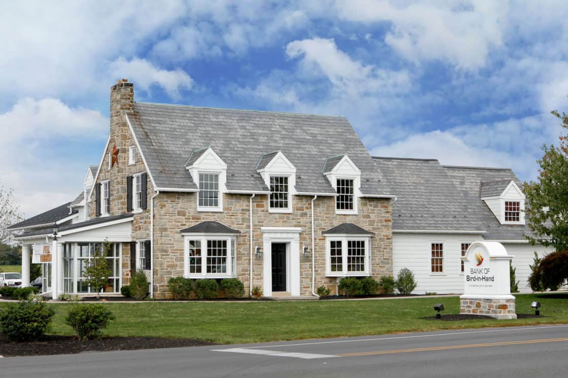 A stone and white-paneled building with multiple dormer windows and a chimney sits on a well-manicured lawn. Reminiscent of a classic bank barn, a sign in front reads “Bank of Bird-in-Hand.” The sky above is partly cloudy.
