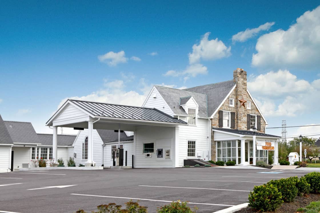 A renovated white building with a stone chimney, drive-thru canopy, and large windows resembles a modern bank barn, sitting in front of an empty parking lot under a blue sky with scattered clouds.