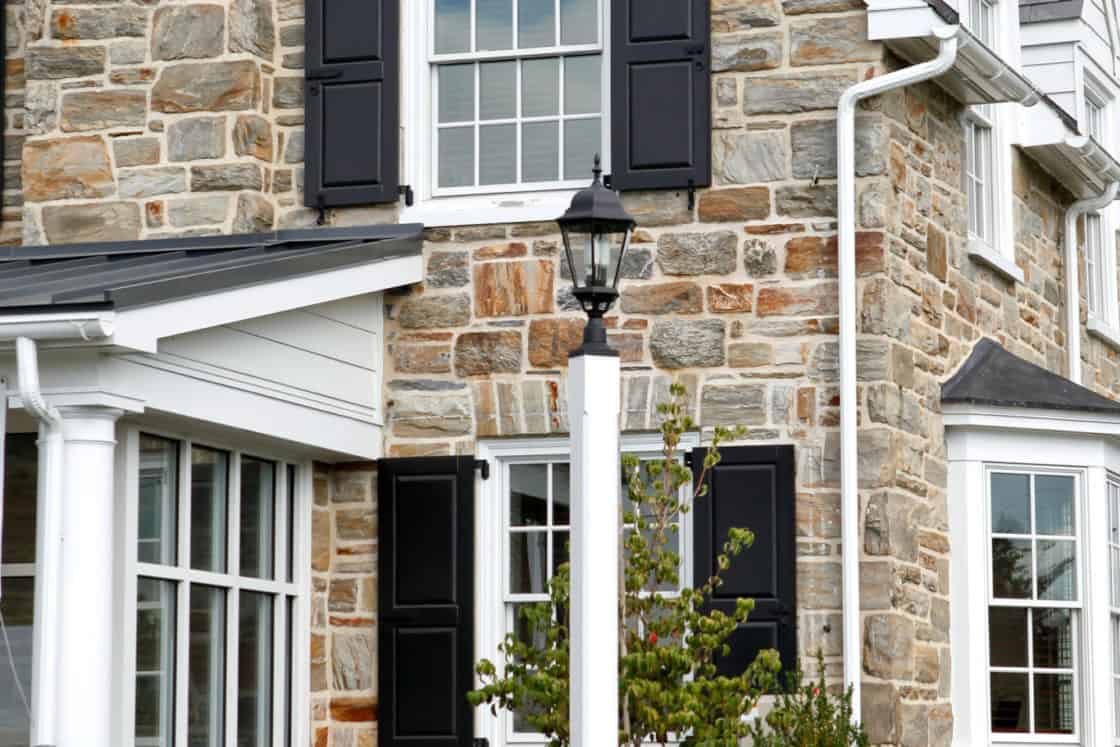 A stone luxury home with black shutters, white trim, and a large star decoration above the window. A black lamp post, green shrubs, and neatly trimmed grass complete the elegant front yard setting.