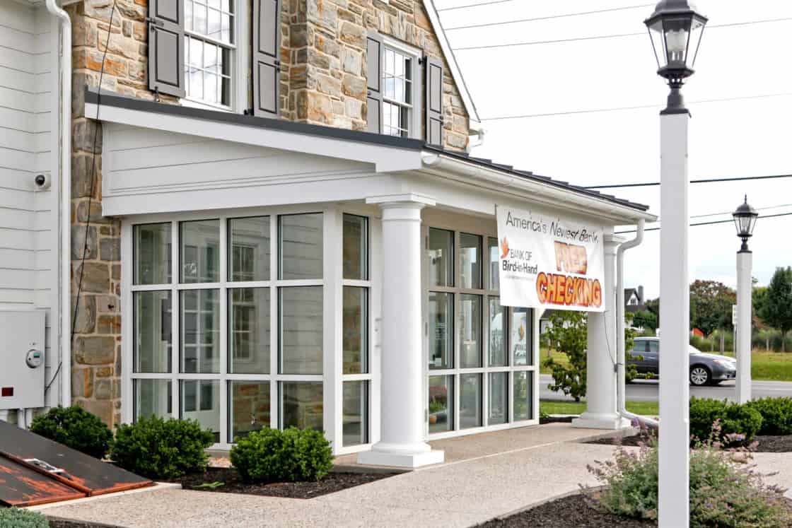 A stone and white building with large windows and a “CHECKING” banner at the entrance, this luxury home is surrounded by shrubs, a paved walkway, a streetlamp, and has a road and greenery in the background.