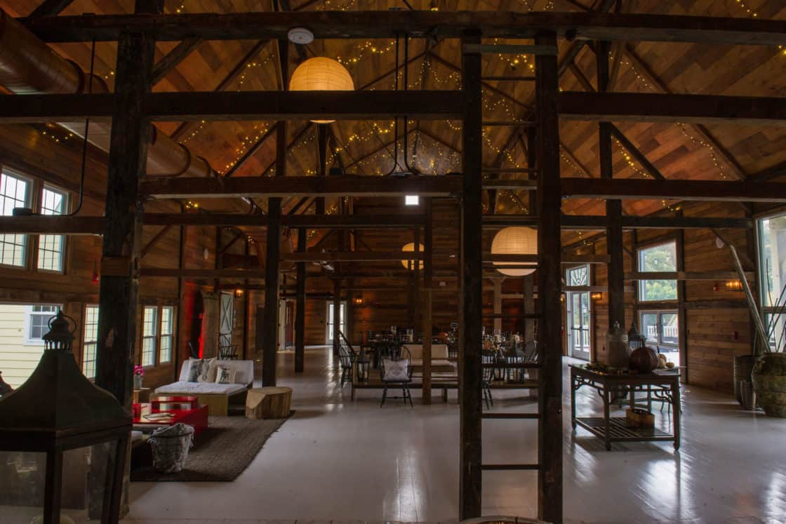 Spacious rustic bank barn interior with wood beams, string lights, large windows, lounge seating on the left, dining tables in the center, and warm lighting creating a cozy, inviting atmosphere.