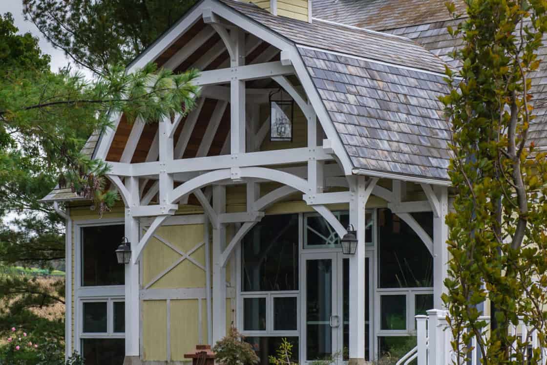 A rustic party barn with a wooden frame, peaked roof, and cupola sits amid landscaped gardens and trees under a cloudy sky. The spacious structure features large windows and a stone walkway leading to its inviting entrance.