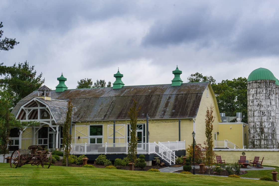 A large yellow party barn with green-roofed cupolas, weathered shingles, and a white ramp sits next to a tall silo with a green dome. There are chairs, neatly trimmed grass, and cloudy skies overhead.