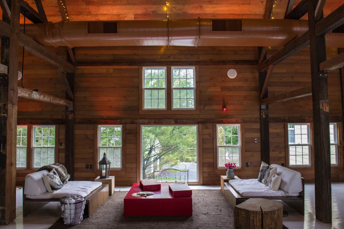 Spacious, rustic living room with wood walls and beams, two white sofas, a red coffee table, and large windows—evoking the charm of a luxury horse barn. Warm lighting and views of greenery create a cozy, inviting atmosphere.
