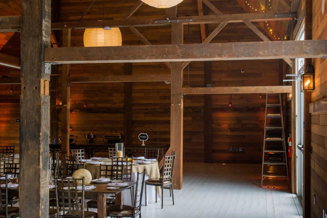 A rustic wooden party barn interior with exposed beams, string lights, hanging paper lanterns, wooden tables and chairs, and a wooden ladder leaning against the wall. Warm natural light brightens the inviting space.