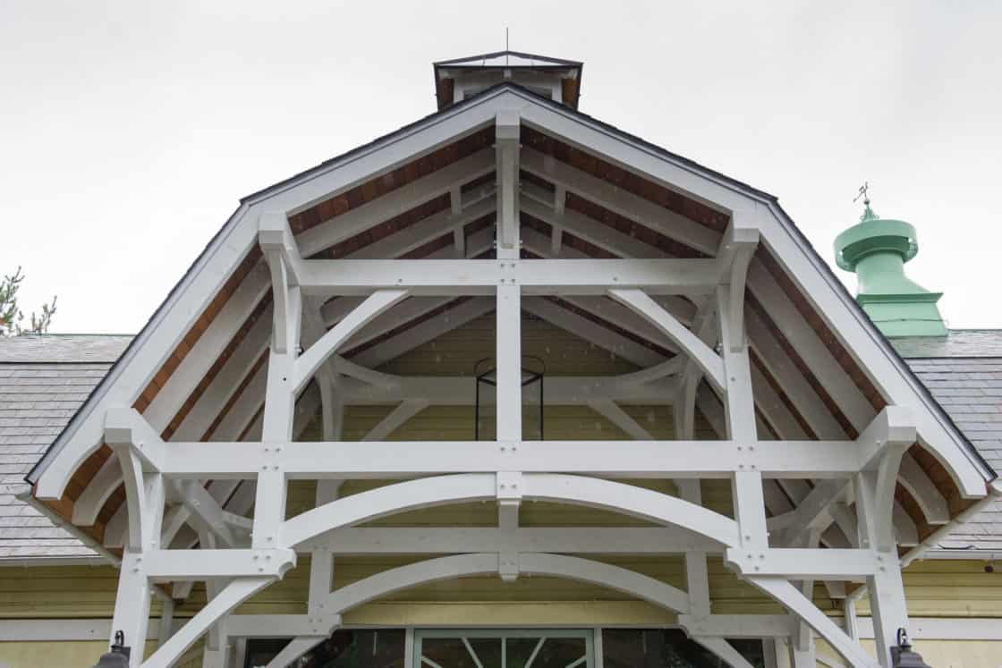 Close-up view of a luxury home entrance featuring a white wooden gable with exposed beams and a peaked roof, set against a cloudy sky.