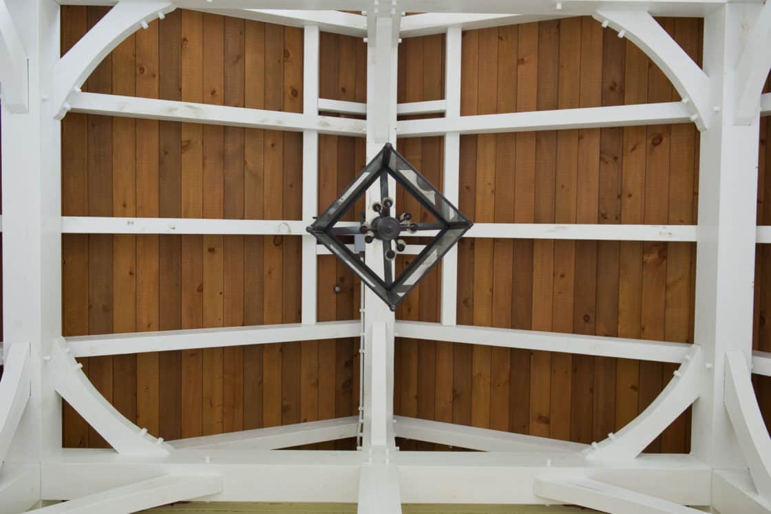 View looking up at a wooden ceiling with white beams arranged in a geometric pattern, reminiscent of a luxury home. A black metal chandelier hangs at the center of the ceiling.