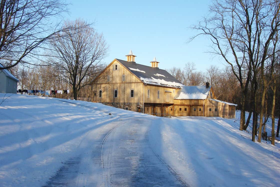 wooden bank barn sitting amidst snow covered landscape surrounded by several trees