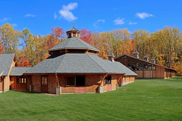 A large, wooden bank barn with a unique octagonal structure in front, surrounded by green grass and autumn trees under a clear blue sky.
