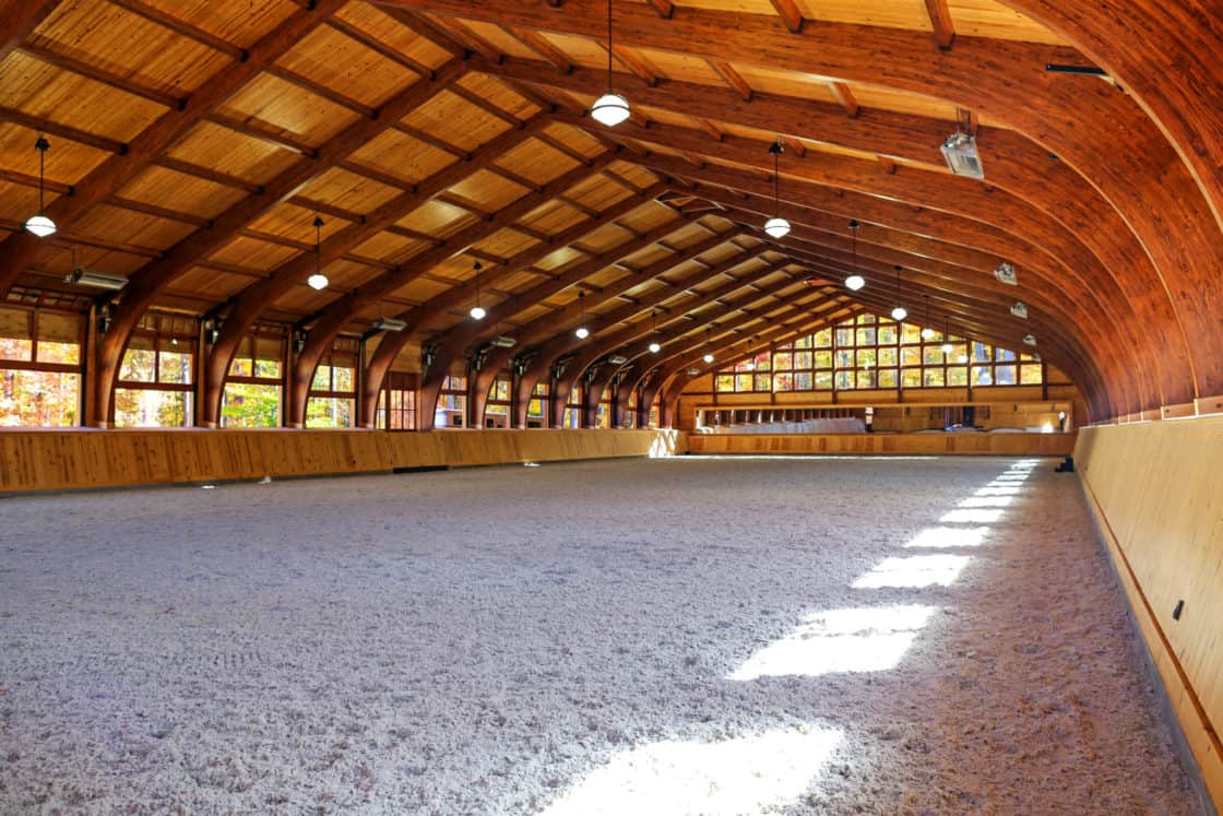 A large indoor riding arena with a high, arched wooden ceiling, like those in a party barn, features windows along the side walls and sunlight streaming onto the sandy ground. The spacious area is open and empty.