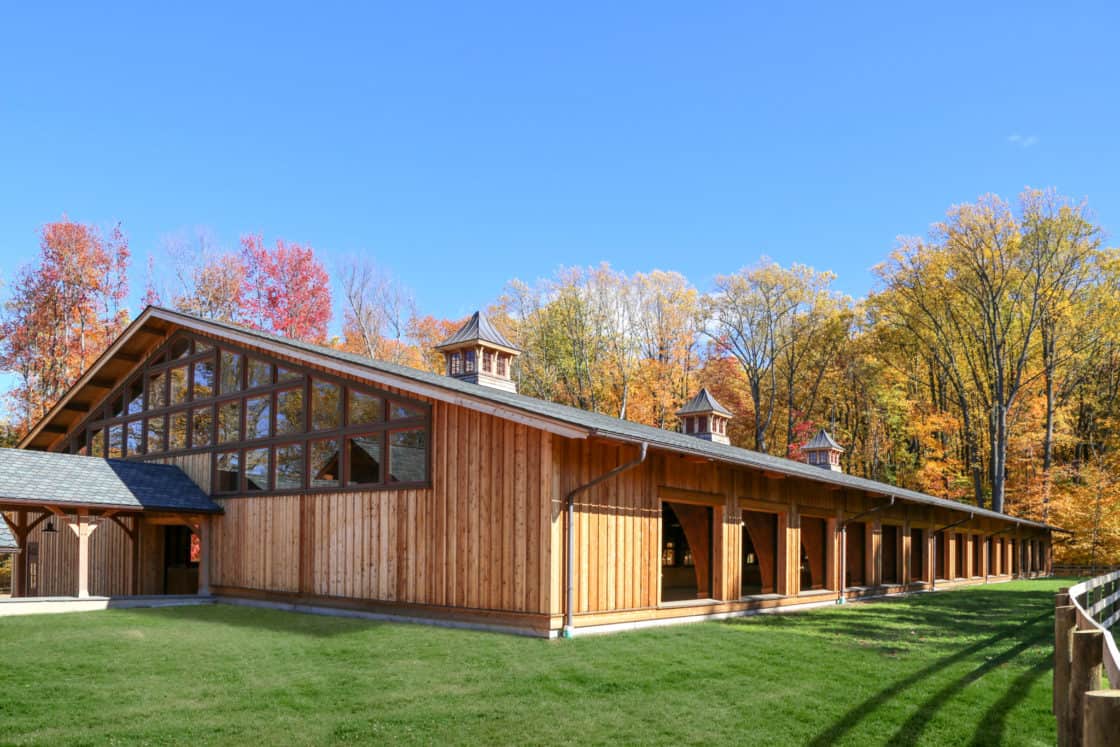 A large wooden luxury horse barn with tall windows and cupolas, surrounded by green grass and colorful autumn trees under a clear blue sky.